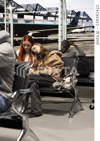 Woman in airport resting head on friend shoulder, feeling upset after delayed flight. Sad girl comforted by sister after losing flight due to airline overbooking, waiting for another plane 139391422