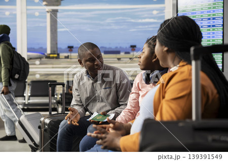 Black parents and teenager seated on bench in bright airport lounge area, surrounded by other travelers near departure gate. Family in good mood waiting for boarding their flight, holiday trip. Black parents and teenager seated on bench in bright airport lounge area, surrounded by other travelers near departure gate. Family in good mood waiting for boarding their flight, holiday trip. 139391549