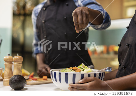 Close up of african american couple preparing food together in their kitchen, adding a pinch of salt to a fresh summer salad bowl. Man and woman enjoying a healthy and organic culinary activity. Close up of african american couple preparing food together in their kitchen, adding a pinch of salt to a fresh summer salad bowl. Man and woman enjoying a healthy and organic culinary activity. 139391678