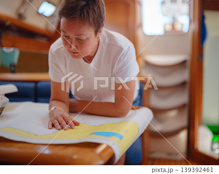 Mature woman sits inside yacht cabin analyzing paper sea chart for route planning. Navigation process involves calculating coordinates and path for upcoming sailing journey from Finike. Mature woman sits inside yacht cabin analyzing paper sea chart for route planning. Navigation process involves calculating coordinates and path for upcoming sailing journey from Finike. 139392461