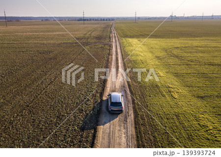 Aerial view of car driving by straight ground road through green fields on sunny blue sky copy space background. Drone photography. Aerial view of car driving by straight ground road through green fields on sunny blue sky copy space background. Drone photography. 139393724