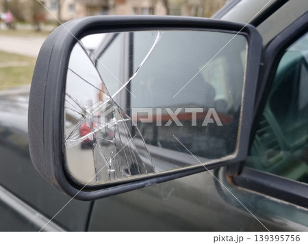 Broken side mirror of a black vehicle showing cracks and damage, with blurred background of parked cars and buildings in a residential area Broken side mirror of a black vehicle showing cracks and damage, with blurred background of parked cars and buildings in a residential area 139395756