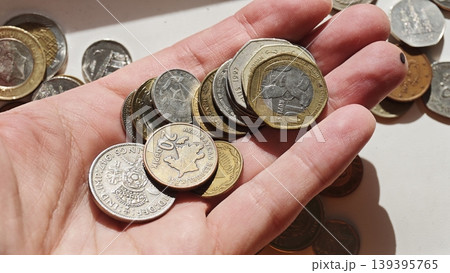 Hand holding assorted coins including various denominations and currencies, with additional coins scattered on a light surface in the background 139395765