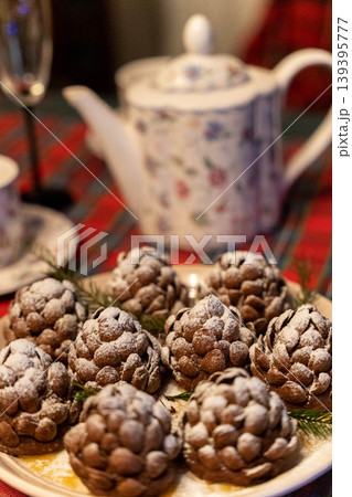 Chocolate pine cone-shaped desserts dusted with powdered sugar on a decorative plate, with a floral teapot and festive tablecloth in the background Chocolate pine cone-shaped desserts dusted with powdered sugar on a decorative plate, with a floral teapot and festive tablecloth in the background 139395777