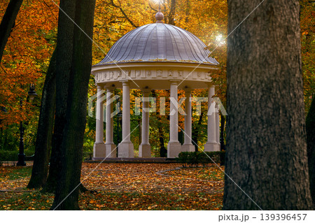 A classical rotunda stands amidst a park ablaze with the vibrant colors of autumn foliage, bathed in soft sunlight 139396457