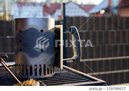 Metal charcoal chimney starter releases smoke on an outdoor grill grate Metal charcoal chimney starter releases smoke on an outdoor grill grate 139396637