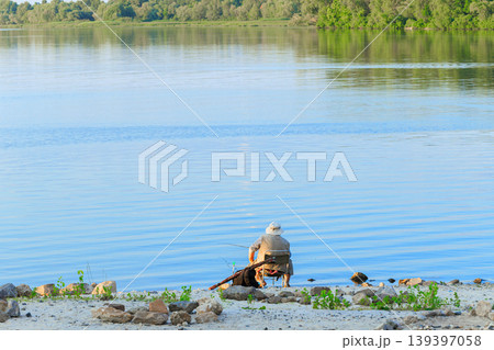 Man sitting on a rocky beach fishing into a calm lake on a sunny day with trees in the background Man sitting on a rocky beach fishing into a calm lake on a sunny day with trees in the background 139397058