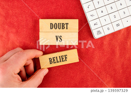 A human hand carefully places a wooden block labeled Belief below stacked blocks reading Doubt vs on a vibrant red textured background. A white calculator rests nearby 139397329