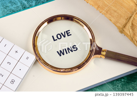 A magnifying glass highlights the words LOVE WINS on a clean white notebook page. A calculator and a rustic paper bundle rest nearby A magnifying glass highlights the words LOVE WINS on a clean white notebook page. A calculator and a rustic paper bundle rest nearby 139397445