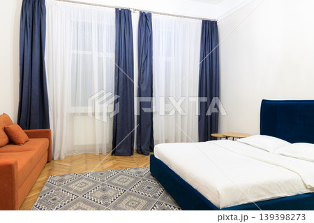 Modern bedroom with orange sofa, blue bed, and patterned rug near window 139398273