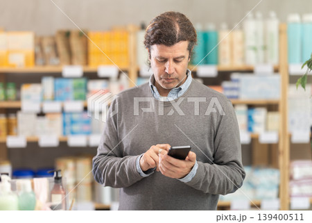 Male shopper searches for necessary goods on the shelves of a supermarket, checking list on her smartphone 139400511