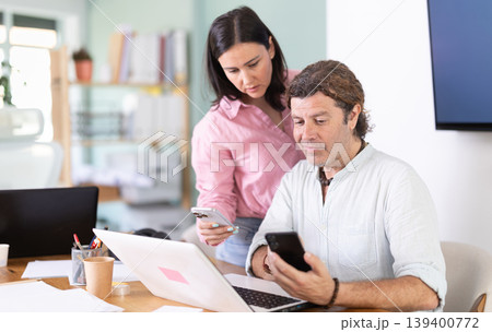 Middle-aged male and female colleagues looking at smartphone at office Middle-aged male and female colleagues looking at smartphone at office 139400772