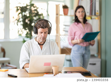 Middle-aged man wearing earphones working with laptop in office Middle-aged man wearing earphones working with laptop in office 139401038