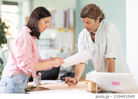 Middle-aged colleagues discussing construction plan leaning over office table Middle-aged colleagues discussing construction plan leaning over office table 139401049