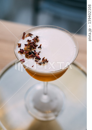 Alcohol beverage. Mixology recipe. Stemmed glass with frothy liquid on silver tray. Restaurant cocktail garnished with dried petals. Alcohol beverage. Mixology recipe. Stemmed glass with frothy liquid on silver tray. Restaurant cocktail garnished with dried petals. 139402108