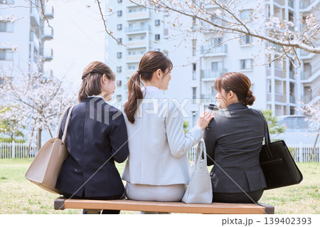 桜の咲く公園のベンチで会話する3人のビジネスウーマン・春の昼休みにリラックスする女性会社員の後ろ姿 139402393
