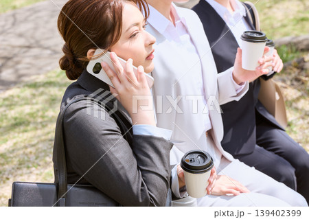 屋外でスマートフォンで通話しながらコーヒーを持つ女性ビジネスパーソンと同僚・休憩中のビジネスシーン 139402399