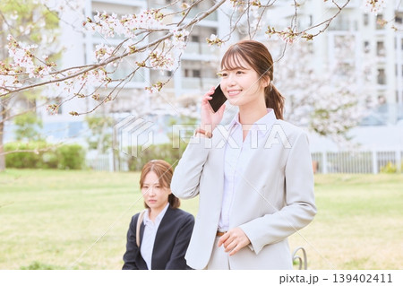 桜の下でスマートフォンで電話をする笑顔の若い女性ビジネスパーソンと背景に佇む同僚・春のビジネスシーン 139402411
