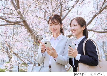 桜の下でスマートフォンを見ながら会話する二人の新入社員・春のビジネスウーマン 桜の下でスマートフォンを見ながら会話する二人の新入社員・春のビジネスウーマン 139402526