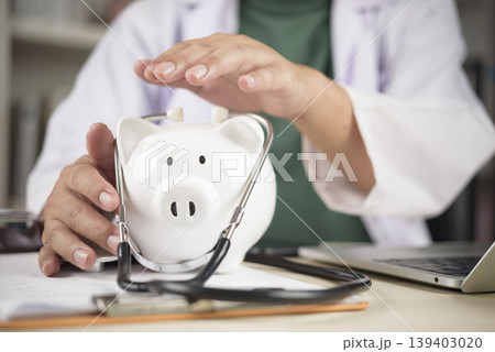 Doctor hands protecting white piggy bank with stethoscope. Concept of medical insurance coverage, healthcare cost safety, and financial security for health. 139403020