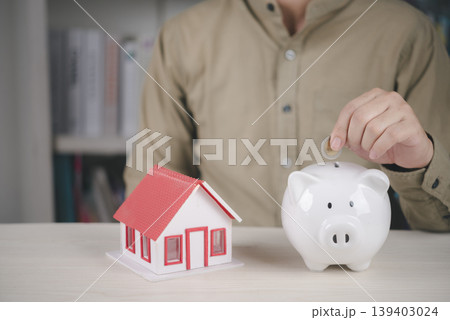 Man putting coin into white piggy bank near house model. Concept of saving money for buying new home, real estate investment, and mortgage financial planning. 139403024