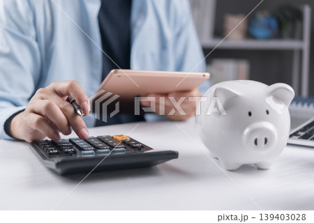 Man using calculator and holding digital tablet for financial planning. Budgeting, tax return calculation and savings concept with white piggy bank on desk. 139403028