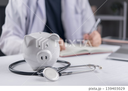 Piggy bank with stethoscope on doctor desk. Health insurance, medical cost and healthcare savings concept. Doctor writing medical prescription in background. Piggy bank with stethoscope on doctor desk. Health insurance, medical cost and healthcare savings concept. Doctor writing medical prescription in background. 139403029