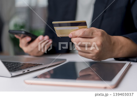 Close-up of hands holding a gold credit card and using a smartphone for online shopping. E-commerce concept with laptop and tablet on the desk. Close-up of hands holding a gold credit card and using a smartphone for online shopping. E-commerce concept with laptop and tablet on the desk. 139403037