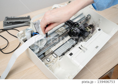 A close-up of a technician's hand using a screwdriver to repair an open inkjet printer, internal carriage, ribbon cable and components. Maintenance, repair, troubleshooting and small electronics work. A close-up of a technician's hand using a screwdriver to repair an open inkjet printer, internal carriage, ribbon cable and components. Maintenance, repair, troubleshooting and small electronics work. 139406753