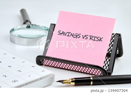 Pink sticky note: SAVINGS VS RISK in holder. Calculator, magnifier, pen on white. Financial planning theme 139407304