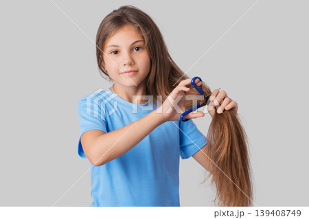 Small apple-cheeked girl in a blue t-shirt going to cut off her long hair with blue scissors against a grey background Small apple-cheeked girl in a blue t-shirt going to cut off her long hair with blue scissors against a grey background 139408749