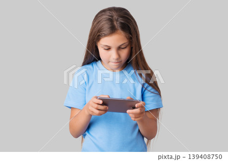 Small concerned girl playing games on smartphone. Young school girl in blue t-shirt attentively looking to her modern device against a grey background 139408750