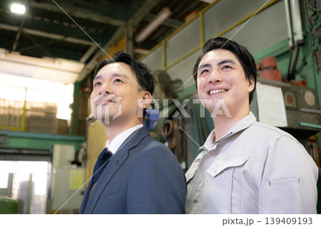 工場で未来を笑顔の横顔で見つめる、見上げるビジネスマンと作業員と2人チームの信頼関係　信金営業や商工 139409193
