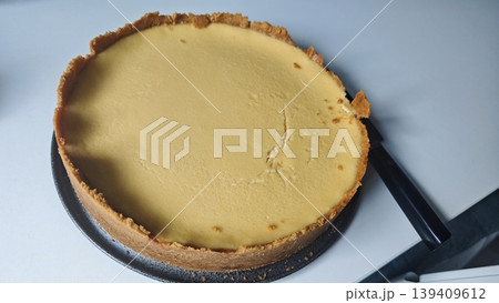 Round cheesecake with a golden crust placed on a white surface, a black knife positioned beside it ready for slicing, showcasing its smooth texture and creamy filling 139409612