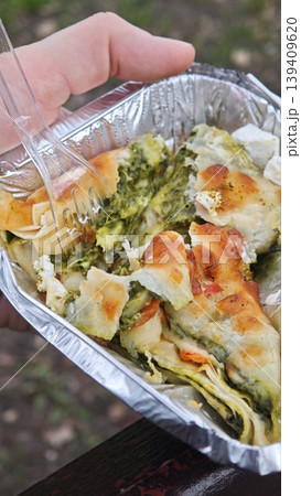 Hand grips a foil tray containing a baked spinach and cheese pastry, revealing a golden crust and green filling, with an outdoor setting in the background Hand grips a foil tray containing a baked spinach and cheese pastry, revealing a golden crust and green filling, with an outdoor setting in the background 139409620
