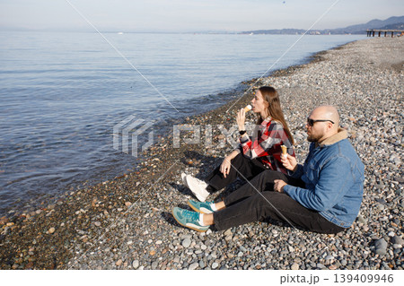 Young couple sitting on beach and eating ice cream Young couple sitting on beach and eating ice cream 139409946