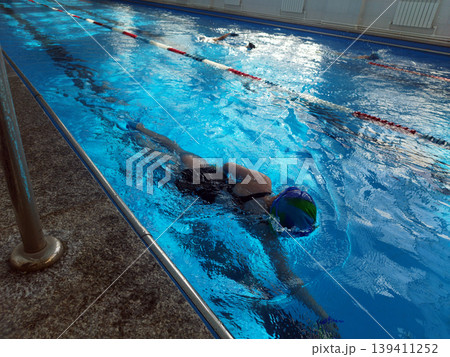 High-angle view of a swimmer in a colorful cap gliding through a bright blue indoor swimming pool. Sunlight reflects off the rippling water as other swimmers exercise in the background lanes 139411252