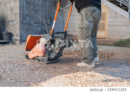 Worker operates a vibratory plate compactor on a gravel surface at a construction site. The machine compacts the ground to create a stable base for paving or structural work. Worker operates a vibratory plate compactor on a gravel surface at a construction site. The machine compacts the ground to create a stable base for paving or structural work. 139411262