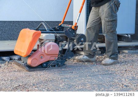 Worker uses a vibratory plate compactor for preparing a base layer. Essential step before paving, decking, or foundation installation. 139411263