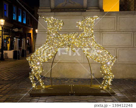 Two glowing golden deer light sculptures facing each other, creating a captivating festive display for holiday season celebration on a cobblestone city street at night 139412248