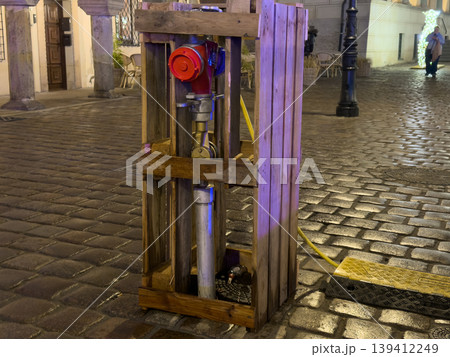 Red fire hydrant securely protected inside a wooden crate on a wet cobblestone street at night, symbolizing urban infrastructure, temporary work, and emergency safety Red fire hydrant securely protected inside a wooden crate on a wet cobblestone street at night, symbolizing urban infrastructure, temporary work, and emergency safety 139412249