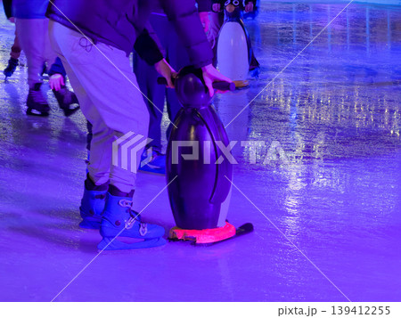 Person ice skating with a penguin skate aid on a purple-lit rink, enjoying winter recreation and learning balance during a festive holiday season activity Person ice skating with a penguin skate aid on a purple-lit rink, enjoying winter recreation and learning balance during a festive holiday season activity 139412255