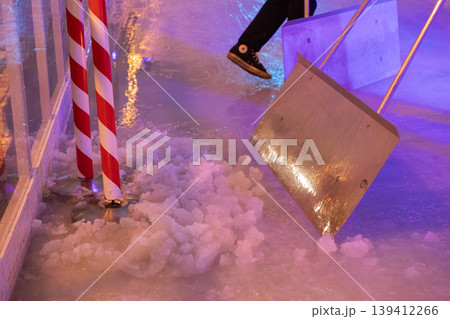 Metal shovel clearing slushy ice on an illuminated rink, near festive candy cane poles, reflecting winter event maintenance and cheerful holiday spirit Metal shovel clearing slushy ice on an illuminated rink, near festive candy cane poles, reflecting winter event maintenance and cheerful holiday spirit 139412266