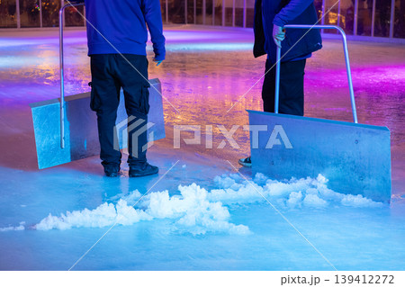Two workers meticulously scrape ice on a vibrant outdoor rink, preparing the surface for public skating with efficient maintenance tools and colorful lighting 139412272