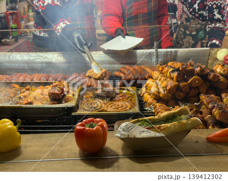 People serving grilled sausages, chicken kebabs, and hot dogs at a lively Christmas market food stall, embodying festive street food culture People serving grilled sausages, chicken kebabs, and hot dogs at a lively Christmas market food stall, embodying festive street food culture 139412302