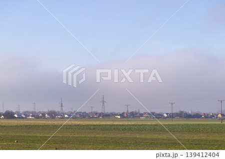 Village houses and prominent electricity pylons emerge from low-lying mist across green fields, highlighting rural energy infrastructure under a clear sky Village houses and prominent electricity pylons emerge from low-lying mist across green fields, highlighting rural energy infrastructure under a clear sky 139412404