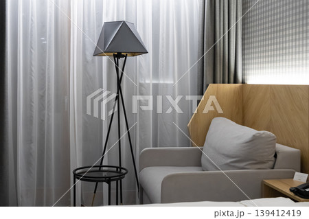 Modern hotel room interior with a striking geometric floor lamp, cozy grey armchair, and contemporary wooden headboard, offering elegant comfort and relaxation 139412419