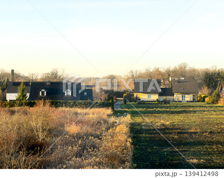 Suburban houses overlooking contrasting fields of dry autumn vegetation and vibrant green land under a bright clear sky, representing peaceful coexistence and seasonal landscape Suburban houses overlooking contrasting fields of dry autumn vegetation and vibrant green land under a bright clear sky, representing peaceful coexistence and seasonal landscape 139412498