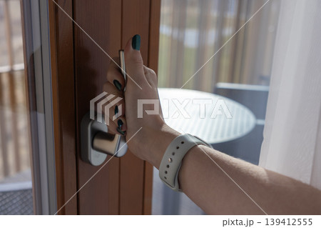 Close-up of a woman's hand with a smartwatch opening a modern wooden balcony door, symbolizing security, access, and the comfort of home or hotel accommodation 139412555