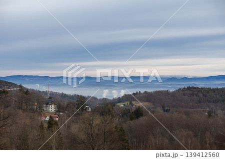 Misty valley landscape with industrial smoke plumes rising, surrounded by bare trees and rural buildings on rolling hills under an overcast winter sky Misty valley landscape with industrial smoke plumes rising, surrounded by bare trees and rural buildings on rolling hills under an overcast winter sky 139412560